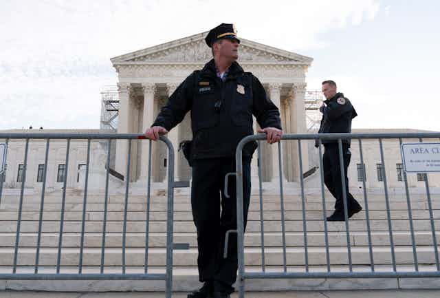 A police officer standing behind a barricade and in front of a large, white columned building.