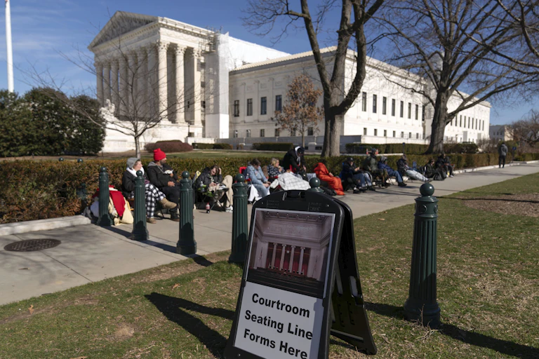 A group of people sit in chairs outside a large white building with columns at its front.
