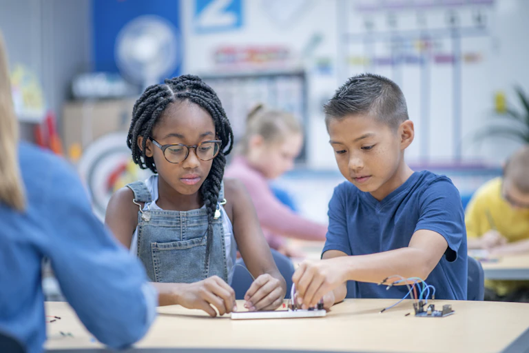 An African American girl works on some sort of electronics project alongside an Asian boy.
