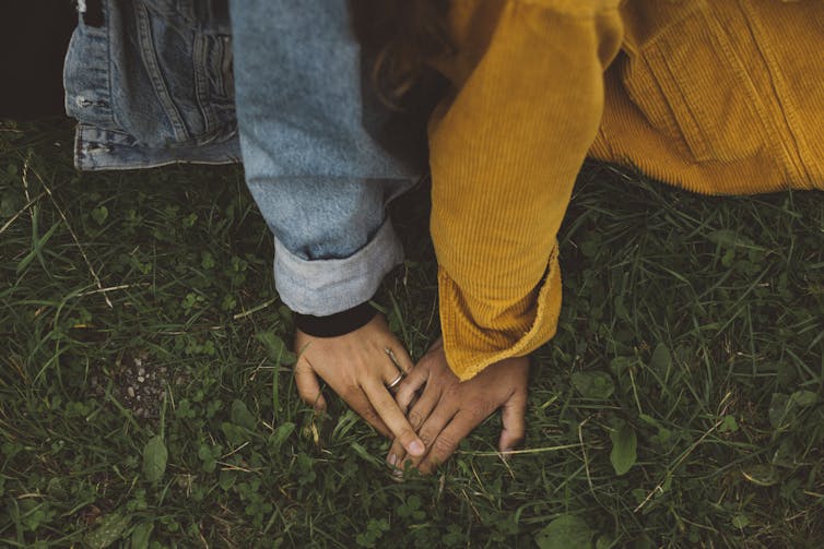Two people sitting back to back on grass, hands loosely intertwined