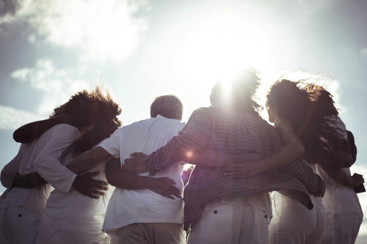 Backs of group of people with their arms links around each other, backlit by the sun