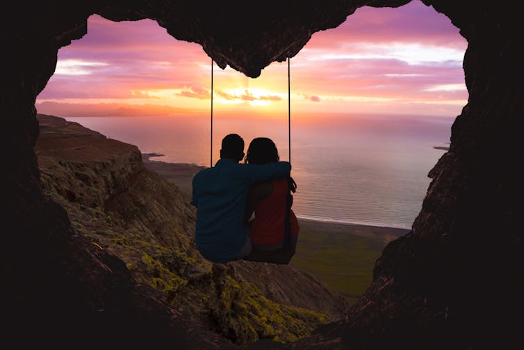 Deux personnes assises sur une balançoire à l'entrée d'une grotte en forme de cœur, regardant la silhouette du soleil se coucher sur l'océan