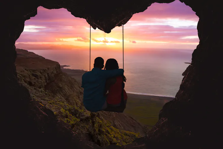 Silhueta de duas pessoas sentadas em um balanço na boca de uma caverna em forma de coração, observando o pôr do sol sobre o oceano