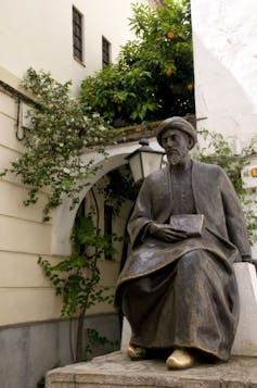 A bronze-colored statue of a man in robes and golden shoes sitting with an open book in his lap, positioned in a sunny courtyard with plants growing behind it.
