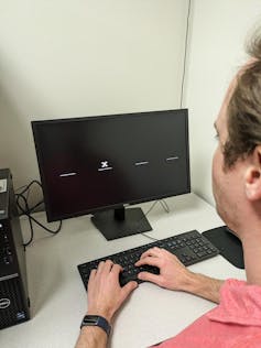 Man in front of computer screen showing four horizontal lines, with an X on one of them.