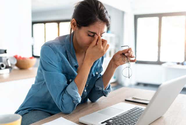 Woman seated at home at laptop pinching the bridge of her nose and looking tired.