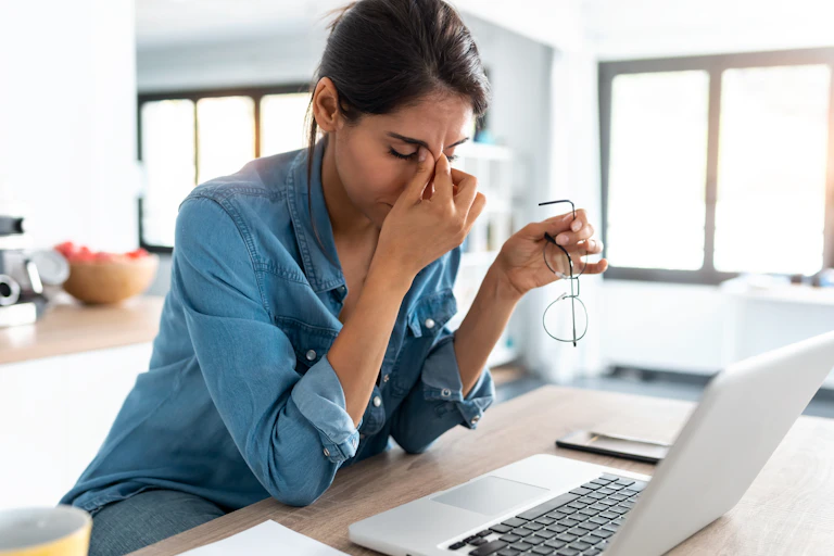 Woman seated at home at laptop pinching the bridge of her nose and looking tired.