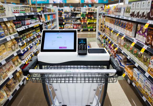 A shopping cart with a tablet and card payment reader built into it sits in the middle of a grocery store aisle