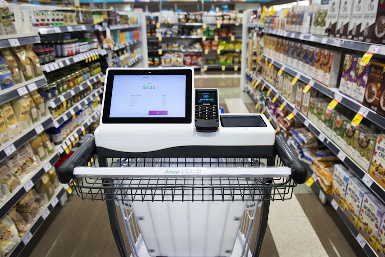 A shopping cart with a tablet and card payment reader built into it sits in the middle of a grocery store aisle