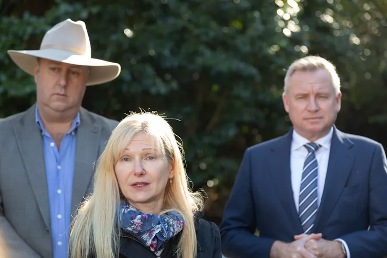 A man in an akubra hat stands behind a short blonde woman. A man in a suit stands on the right