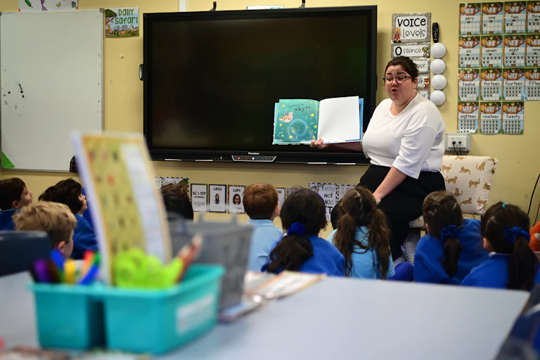Students sit in front of their teacher, who is reading them a book.