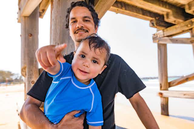 An Aboriginal man holds his toddler son at the beach