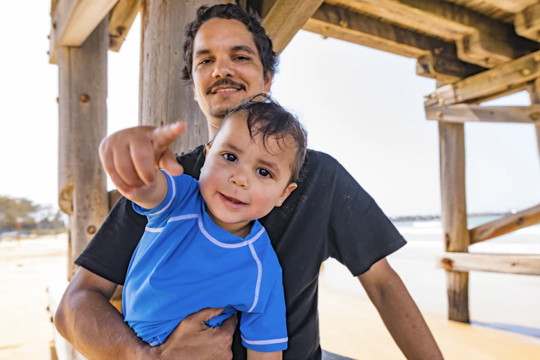 An Aboriginal man holds his toddler son at the beach