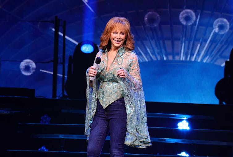 Woman with red hair and silver dress holds microphone and smiles.