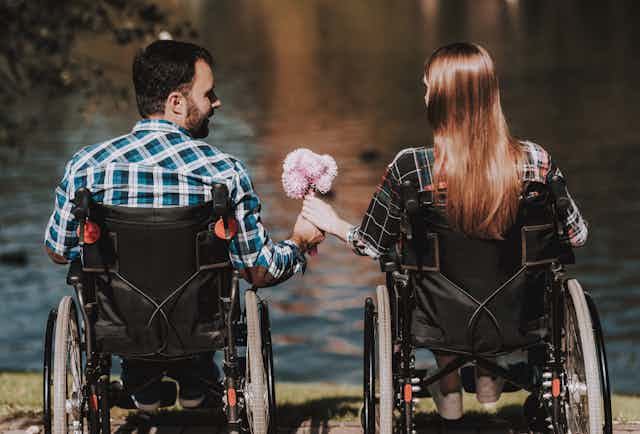 A man and woman in wheelchairs sit in front of a lake. He hands her a bouquet of flowers.