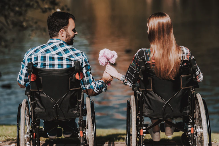 A man and woman in wheelchairs sit in front of a lake. He hands her a bouquet of flowers.