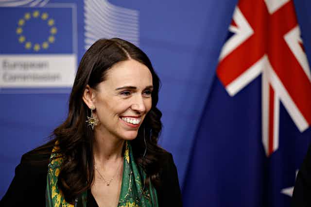 Woman with long dark hair, smiling, smart clothes, against a blue backdrop with EU and NZ flags