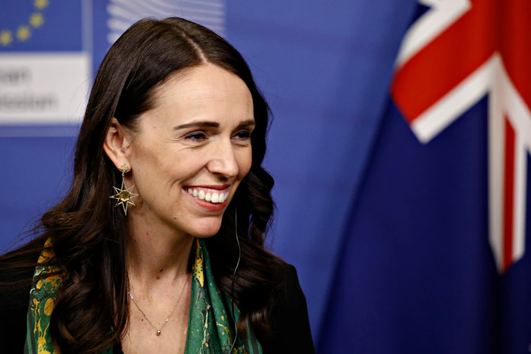 Woman with long dark hair, smiling, smart clothes, against a blue backdrop with EU and NZ flags