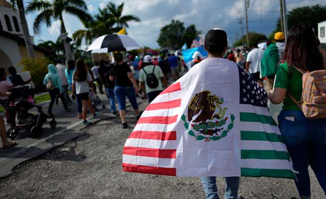 A man walks draped in a mashup of the United States and Mexico flags in a tropical place at the end of a big procession.