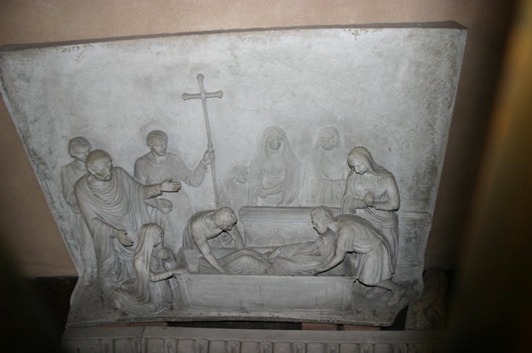 A relief showing a corpse being placed in a coffin as people stand around, one holding a tall crucifix.
