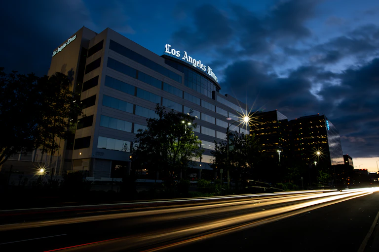 The Los Angeles Times headquarters at night with the streaking headlights of cars driving by in the foreground.