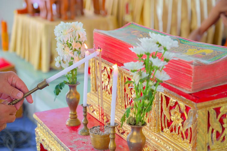 A man lights a candle on an altar, painted red, with white flowers in two vases and incense sticks in a small pot.