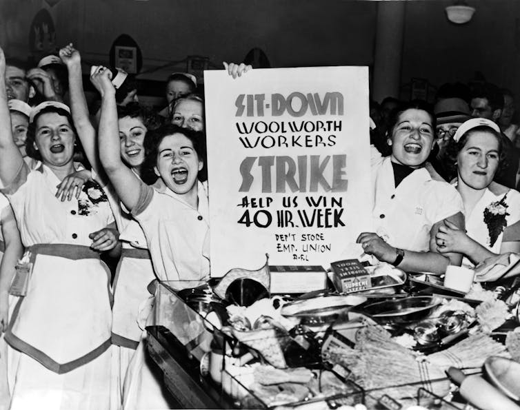 Young women raise their fists and smile. Two of them hold a sign reading 'SIT-DOWN STRIKE - HELP US WIN 40 HOUR WEEK.'