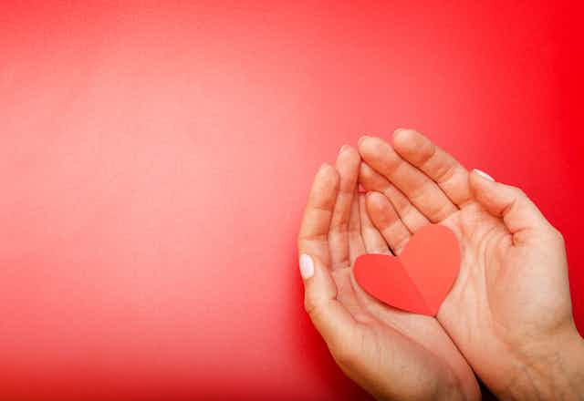 Two hands holding a red paper heart against a red background