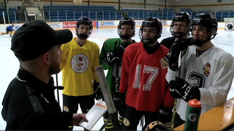 A man in a black shirt and baseball hat talks to a group of teenage hockey players on an ice rink