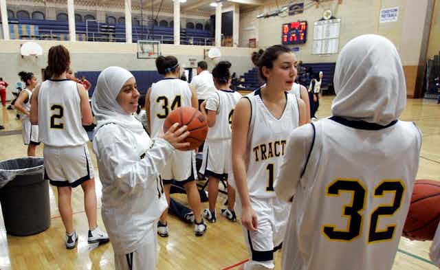 In a school gym, teenage girls wear white basketball uniforms. Several of the girls have their hair covered by a white scarf.
