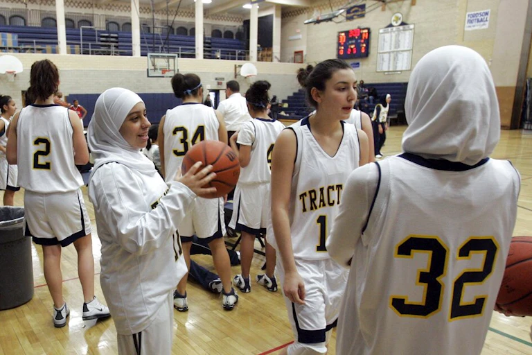 In a school gym, teenage girls wear white basketball uniforms. Several of the girls have their hair covered by a white scarf.