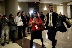 A smiling woman with black hair and glasses claps as she walks down a hallway wearing a lanyard.