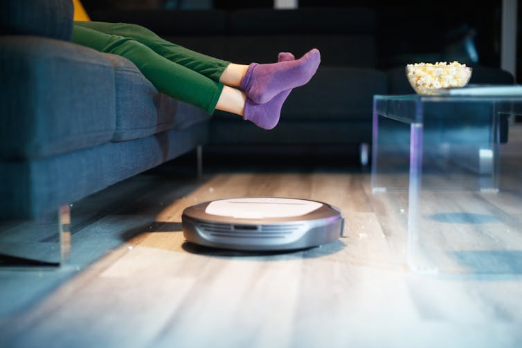 A shiny round vacuum cleaning up popcorn crumbs under a person on sofa wearing purple socks