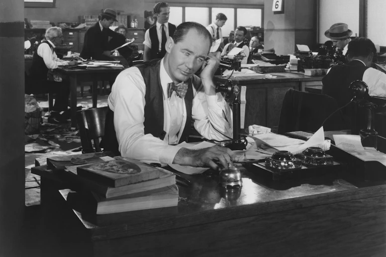 Older worker in bow tie and waist coat sits at desk while talking on the phone.