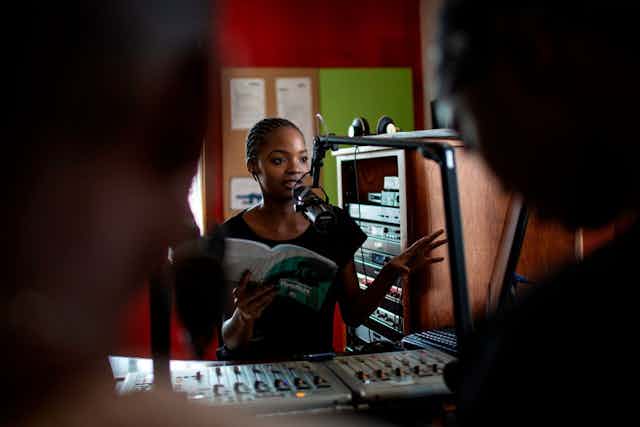 A young woman is seated in a radio station, talking into a microphone, gesturing with one hand while holding a book with the other as two blurred figures watch her in the foreground.