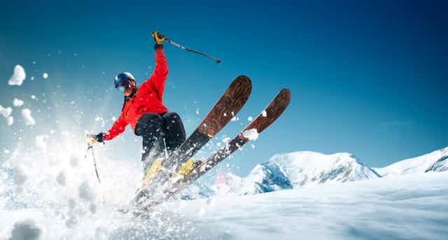 Skiier wearing helmet and red jacket, skis airborn from the slope, snow flying towards camera, blue sky and mountains in background