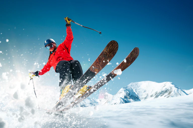 Skiier wearing helmet and red jacket, skis airborn from the slope, snow flying towards camera, blue sky and mountains in background