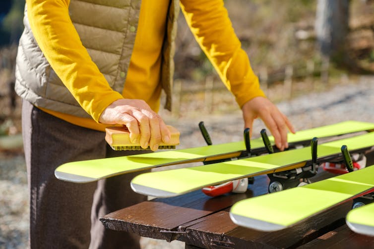 Woman in yellow jackets applies wax to four yellow skis laid out on wooden table