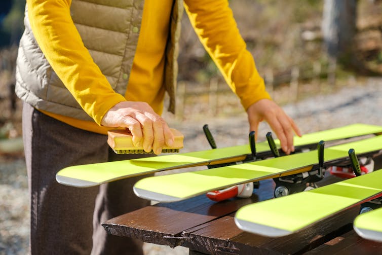 Une femme vêtue d’une veste jaune applique de la cire sur quatre skis jaunes posés sur une table en bois.