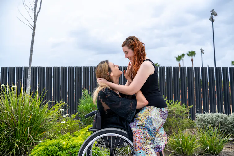 A woman sits on the lap of another woman in a wheelchair. They look at each other lovingly.