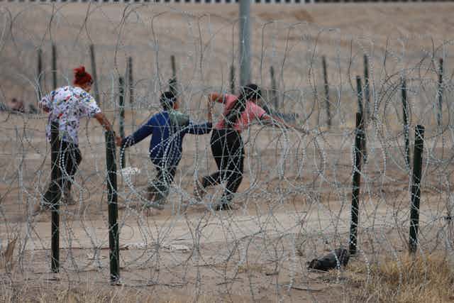 Three people are seen holding hands and running through barbed wire in an arid landscape.