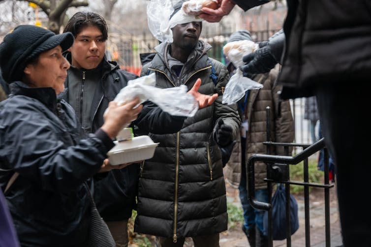 People wearing dark clothing and jackets reach for and hold bags of bread.