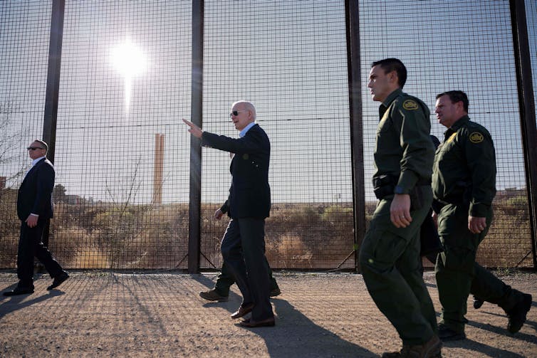 Joe Biden wears dark sunglasses and a suit and walks, in front of men in green uniforms, along a large fence. The sun shines through it.