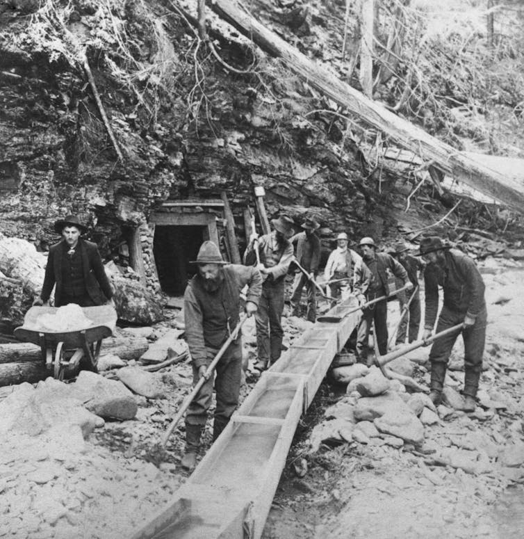 Men wield shovels and wheelbarrows next to a small timbered opening in a hillside.