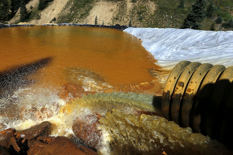 Bright orange water flows from a pipe into a plastic-lined settling pond.
