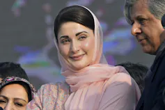 A dark-haired woman in a pale pink sari and head scarf smiles as she listens to someone speak.