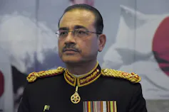 A thin balding man in a military uniform wearing glasses looks solemn at an event.
