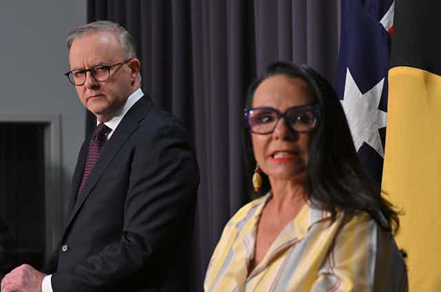 Anthony Albanese stares at the camera behind Linda Burney, who's speaking to the media