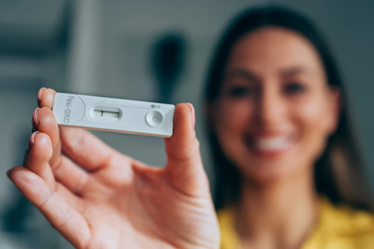 A smiling woman, holding a rapid home test device, shows her negative results.