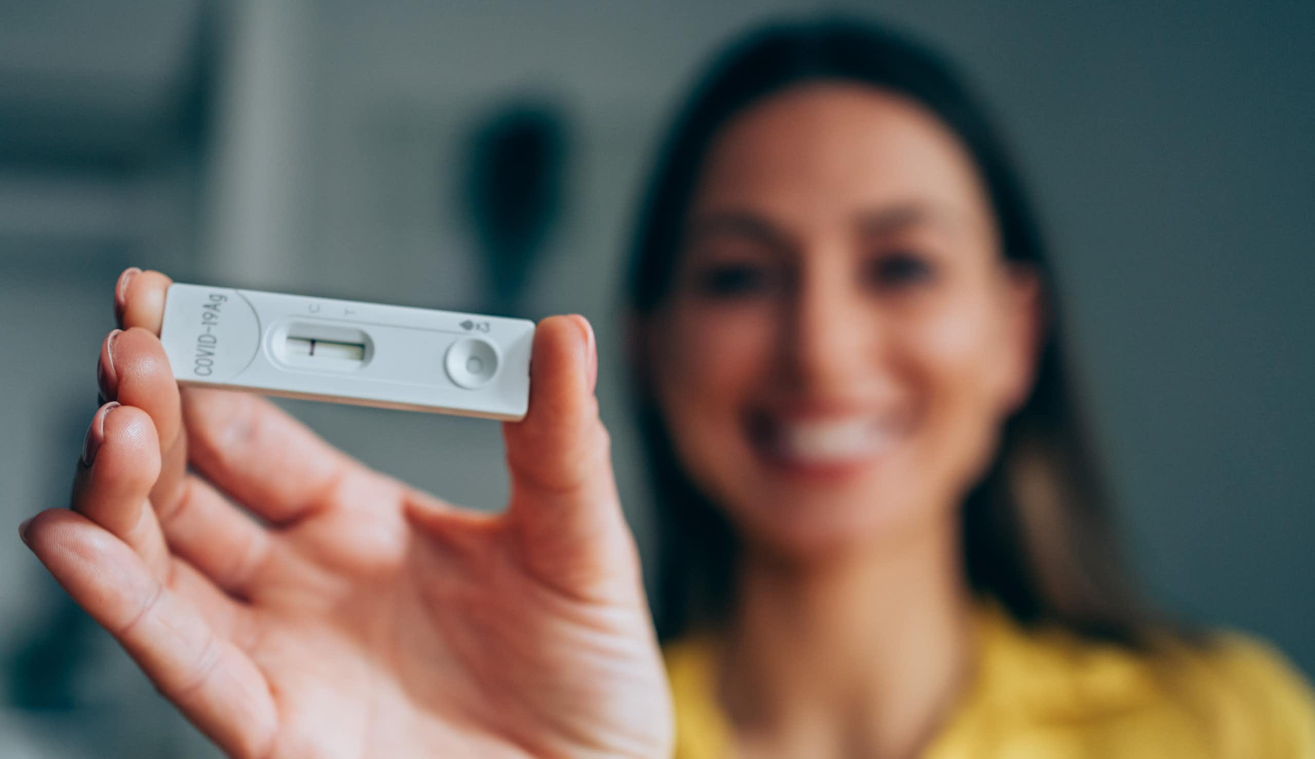 A smiling woman, holding a rapid home test device, shows her negative results.